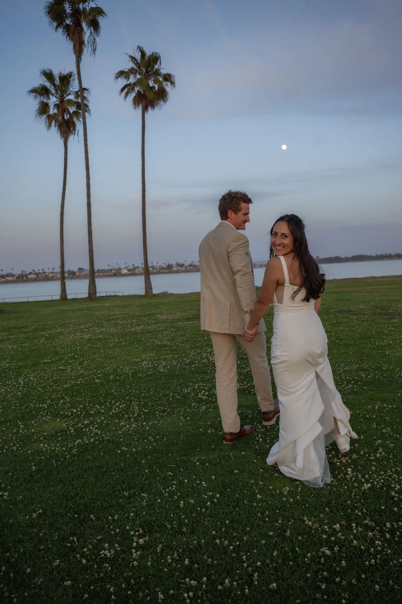 Bride and groom hold hands at sunset among the palms by the bay, with a rising moon