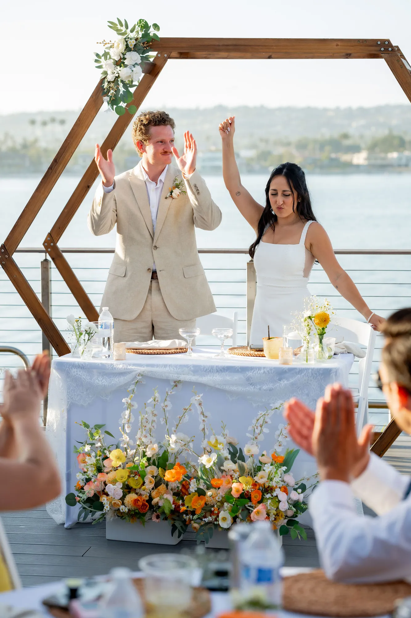 Newlyweds cheer at their sweetheart table beneath a hexagon floral arch overlooking the bay