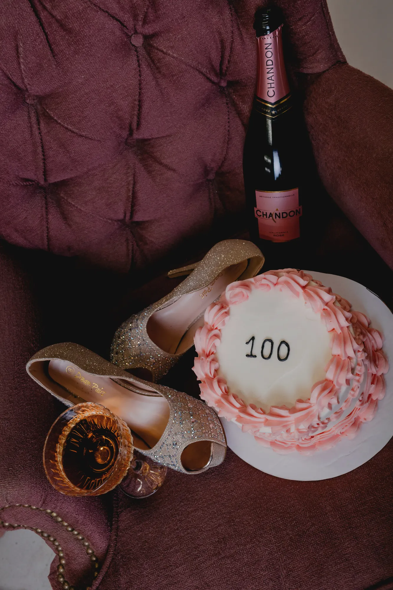 Gold heels, rosé champagne, and a pink 100 cake on a velvet chair at Jen's milestone shoot